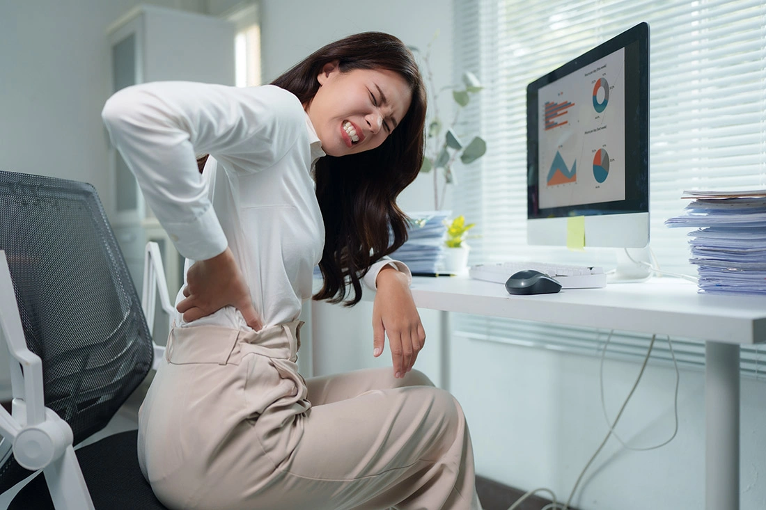Office worker sitting at a desk and holding their lower back in discomfort, illustrating work‑related back pain.