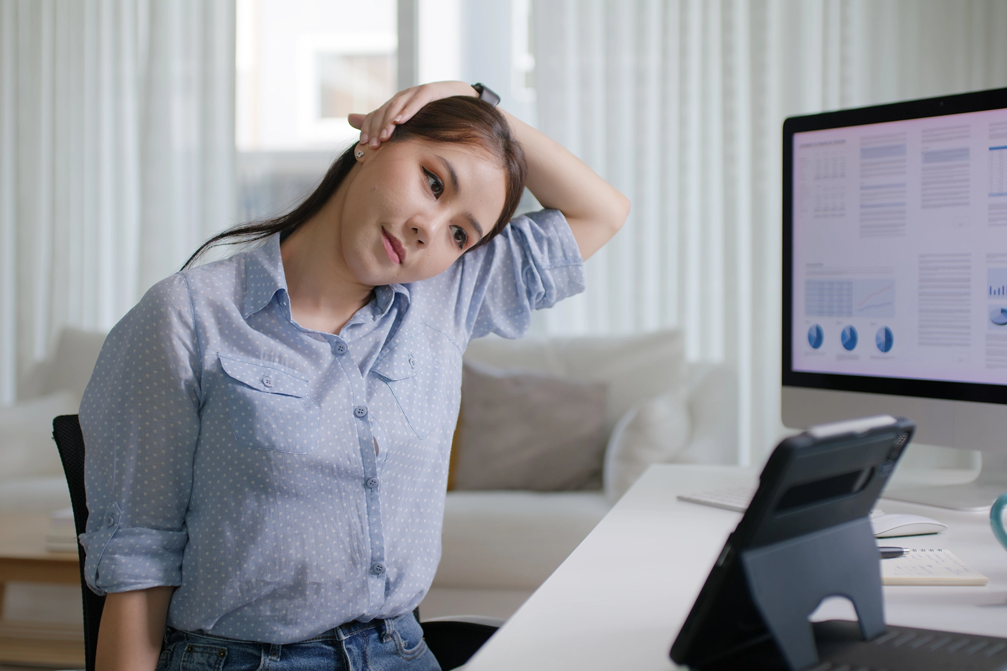 Asian woman at her desk performing neck exercises, stretching to relieve tension and improve posture while working.