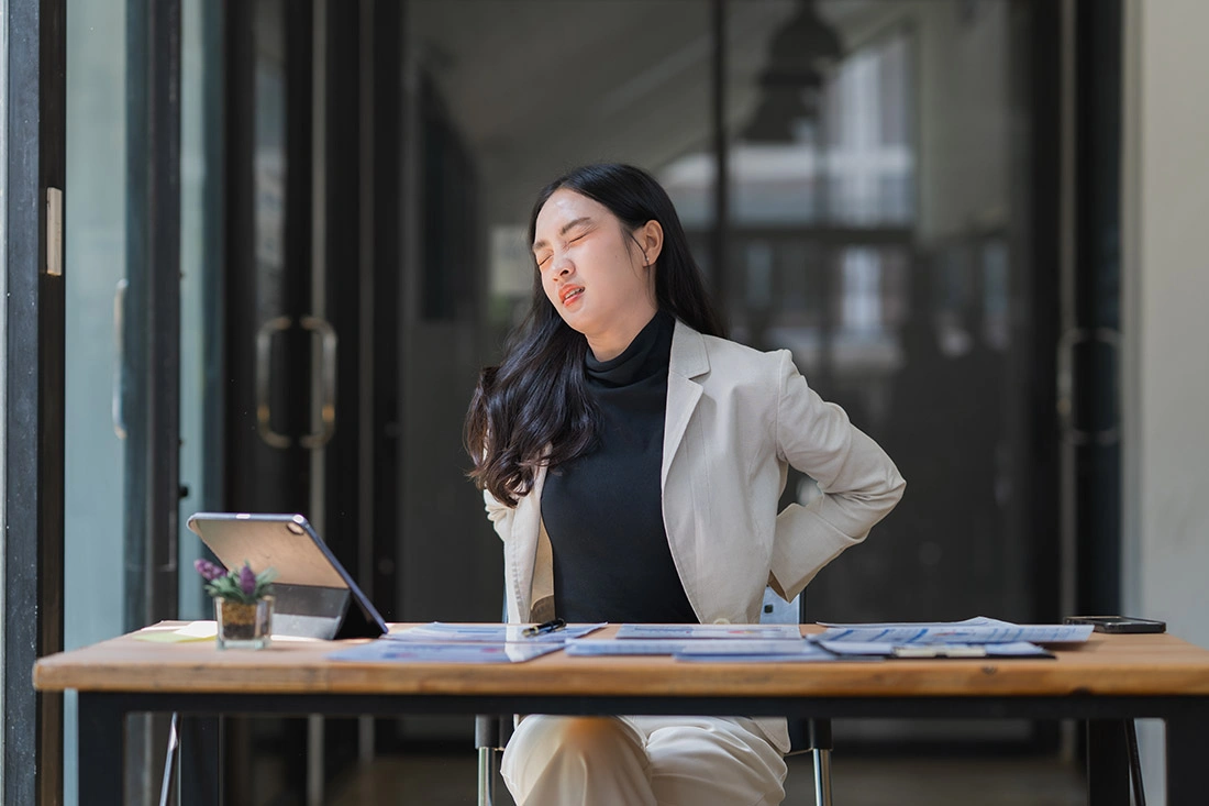 Asian woman grimacing and holding her lower back in pain, illustrating discomfort from lower back strain or injury.