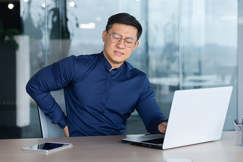Man sitting at an office desk holding his lower back in discomfort.