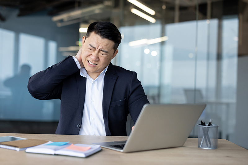 Asian professional at desk showing signs of neck strain with hand on neck, illustrating neck pain.