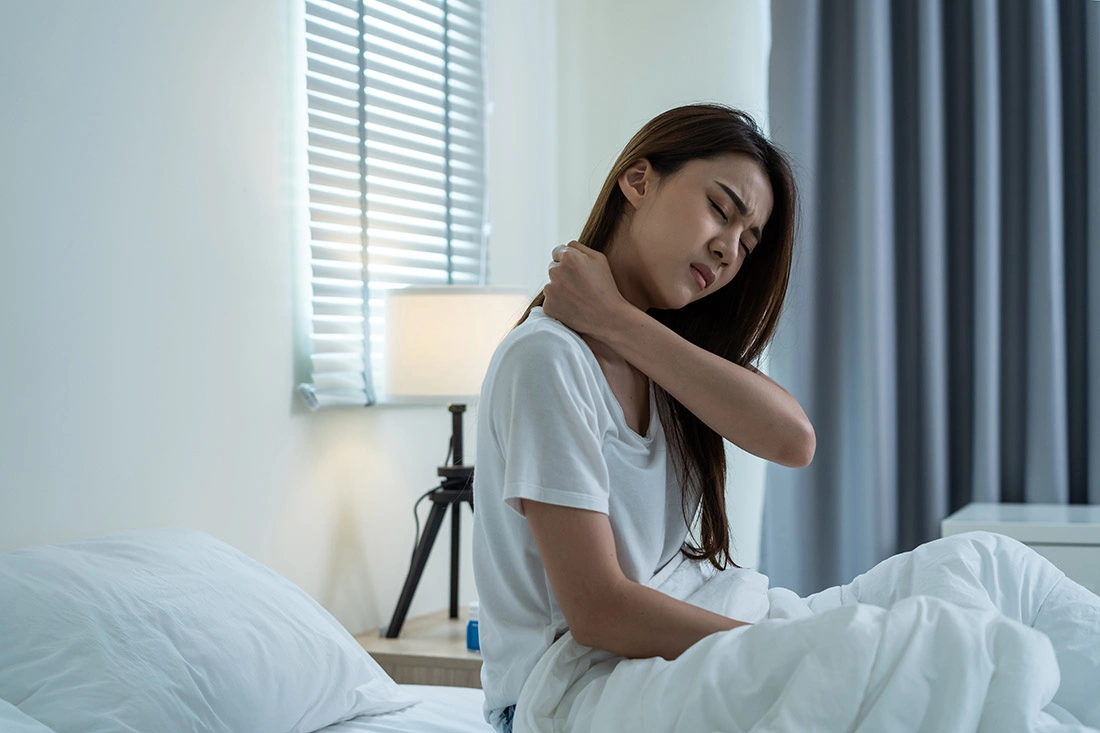 Asian woman sitting on her bed and holding her neck in pain.